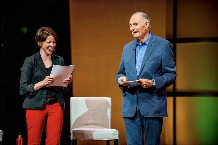 Bernadette Woods Placky laughing on stage alongside Alan Alda, both holding scripts
